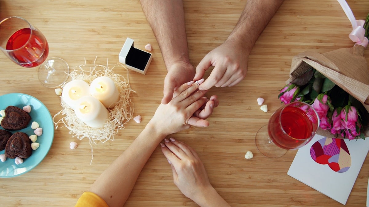 Guy is putting beautiful engagement ring on girl's finger at table in restaurant on Valentine's day then couple is holding hands enjoying dinner. People and relations concept.